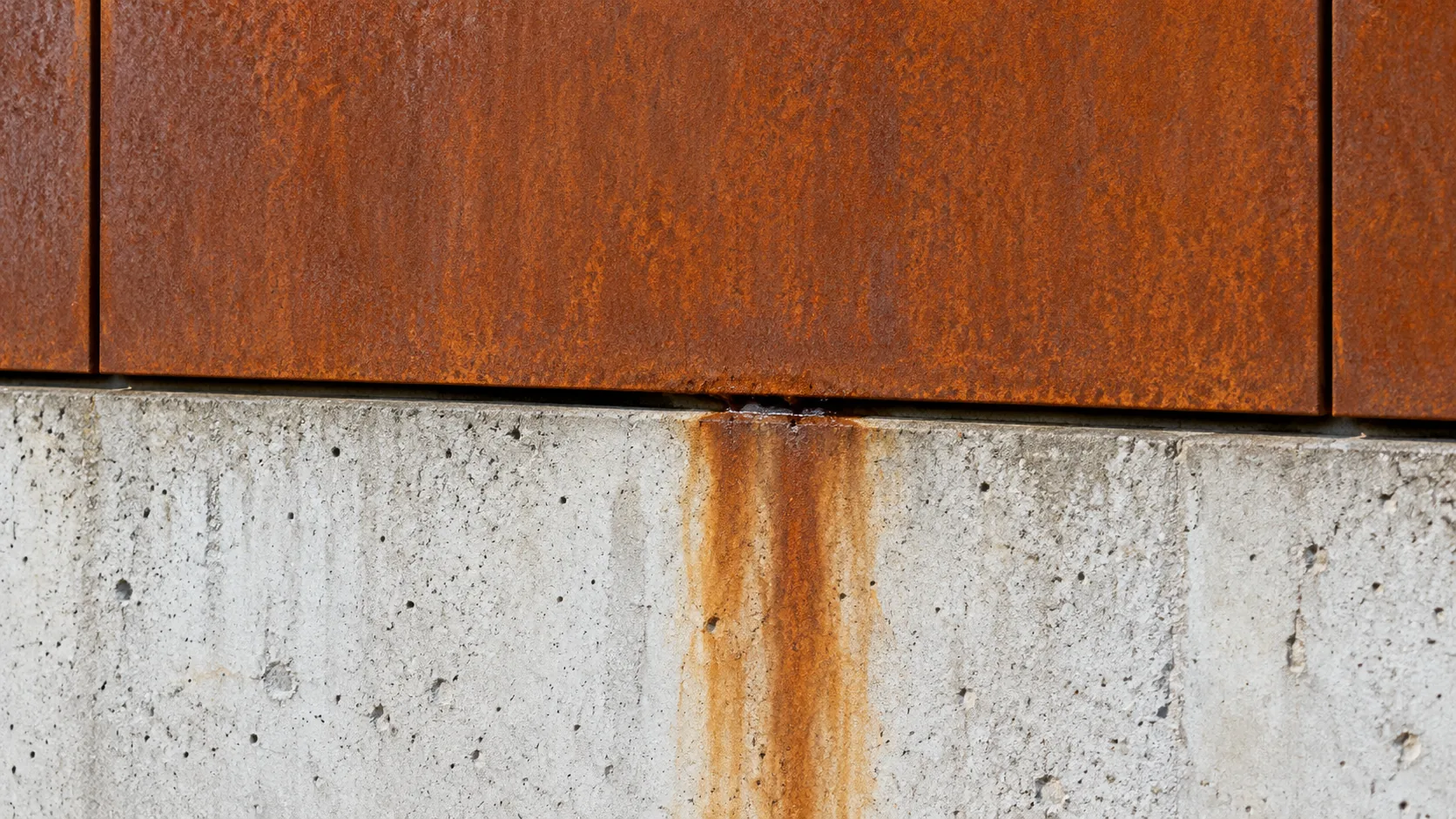 Orange-brown weathering-steel panel above pale concrete, with a brown stain running down from the drip.