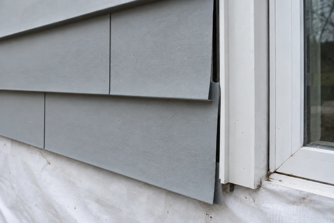 Close-up of horizontal light-gray steel lap siding courses meeting a vertical white J-channel trim receiver at a window jamb over a white weather barrier, showing the uncaulked expansion gap and staggered lap joint.